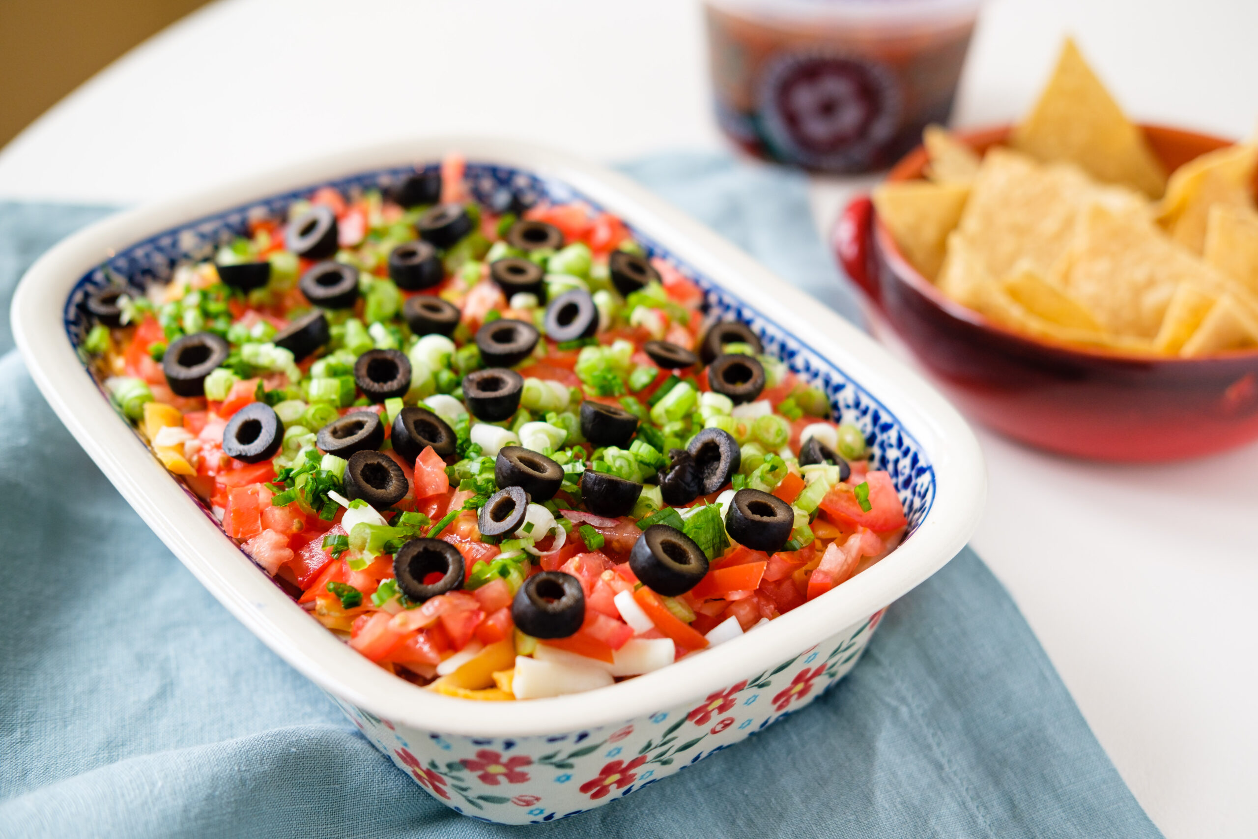 A casserole on a table, with a bowl of tortilla chips and container of salsa in the background.