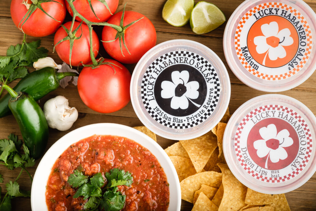 Three Lucky Habanero containers, a bowl of salsa, and chips and produce scattered on a wooden table.
